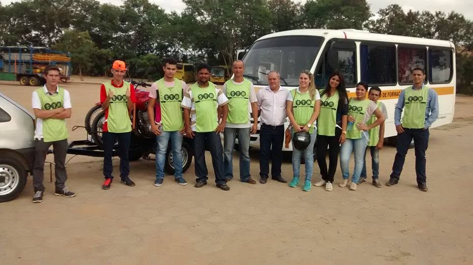 Doze alunos aprovados de colete verde, em frente ao micro-ônibus e à moto da autoescola