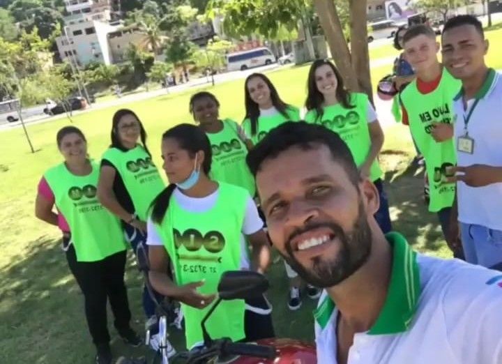 Instrutor e turma de alunas e alunos de colete verde, reunidos em uma área verde antes da aula de moto